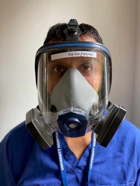 A doctor wearing scrubs and a full-face respirator, which has a nametape reading 'Sanjay Agrawal' stuck to it