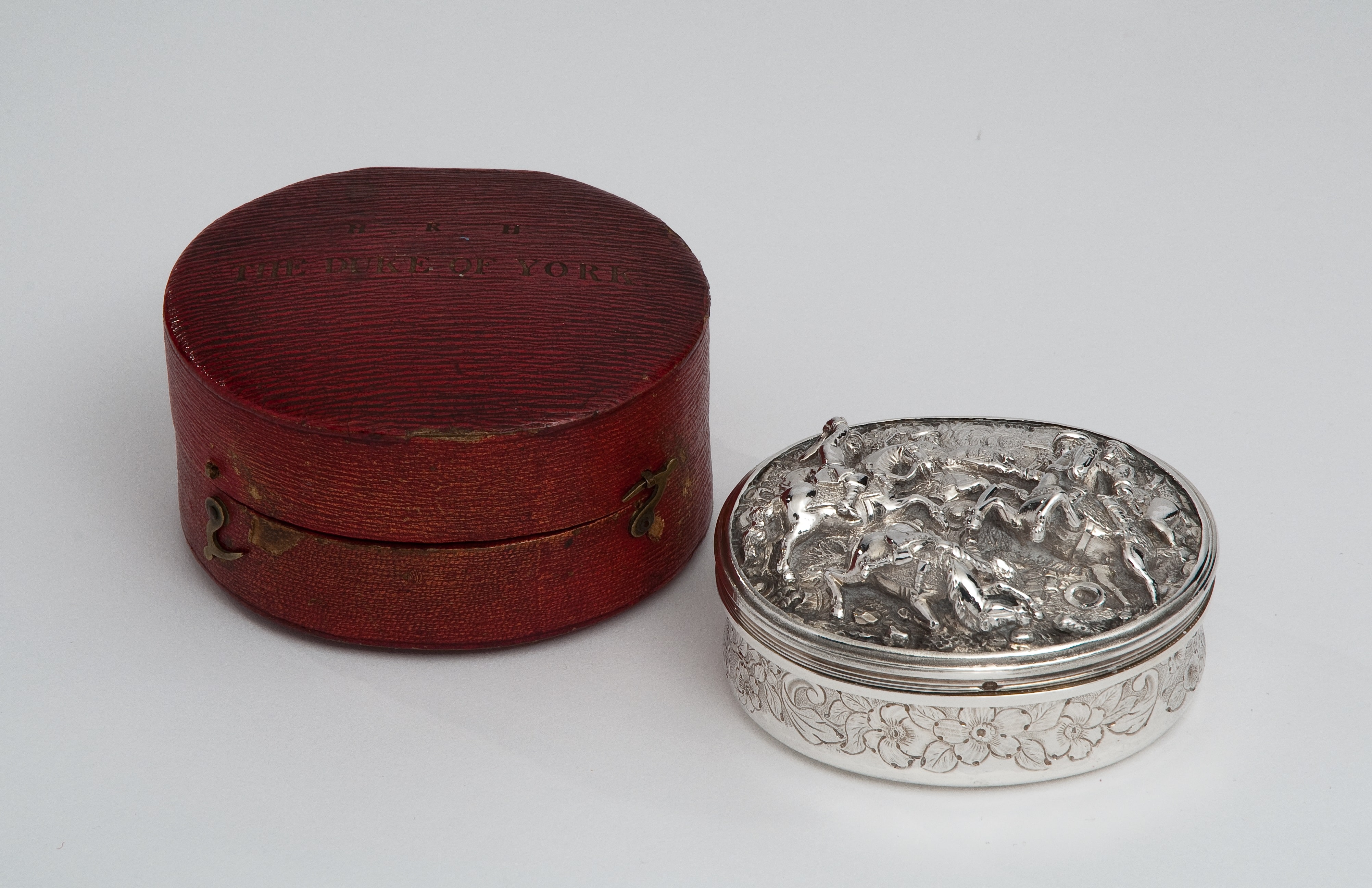 Silver circular box with engraved design on top, with red leather circular case to left