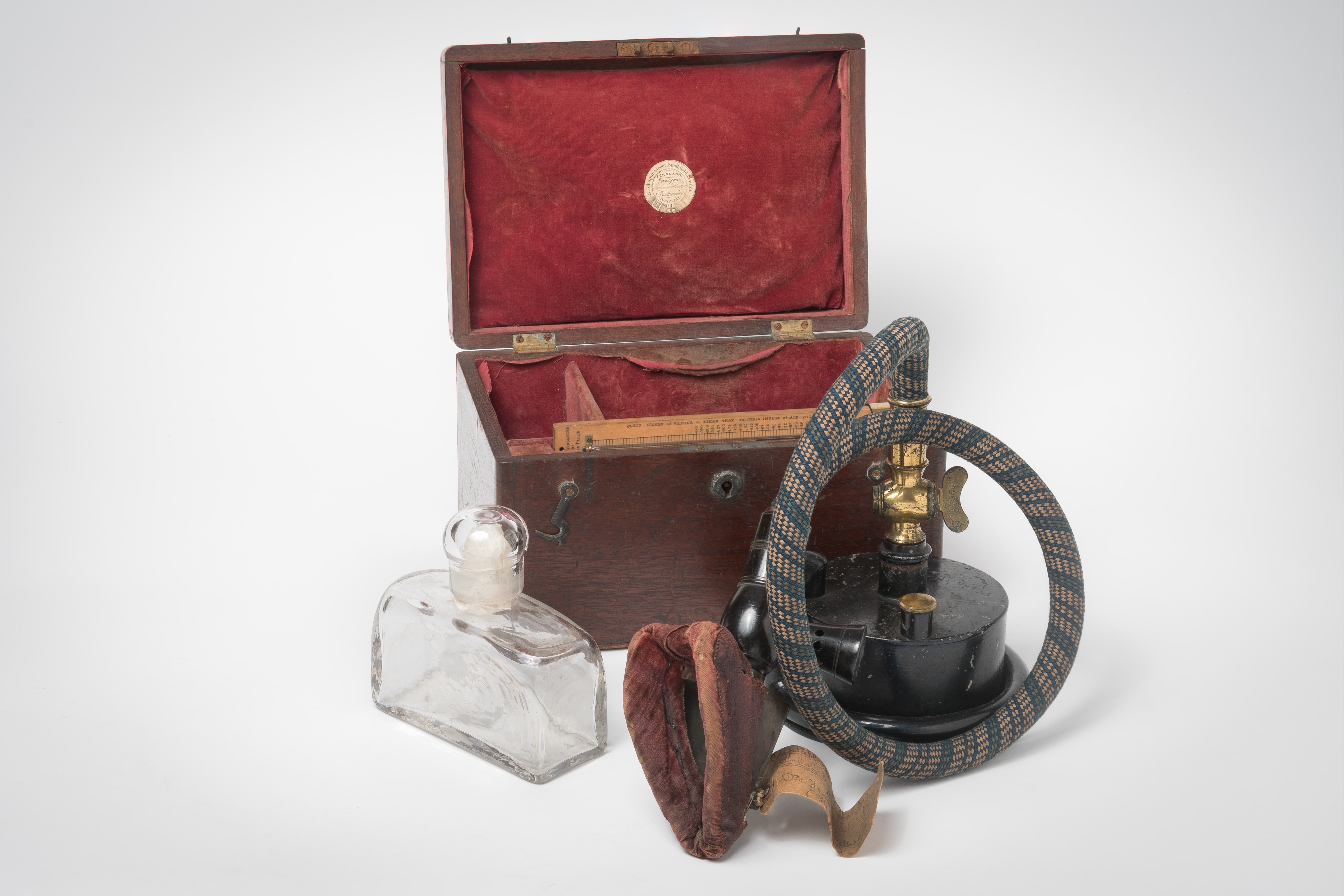 Wooden box with red linking, propped open. In front, a glass flask, and a anaesthesia facemask made of red velvet attached to a small metal cannister