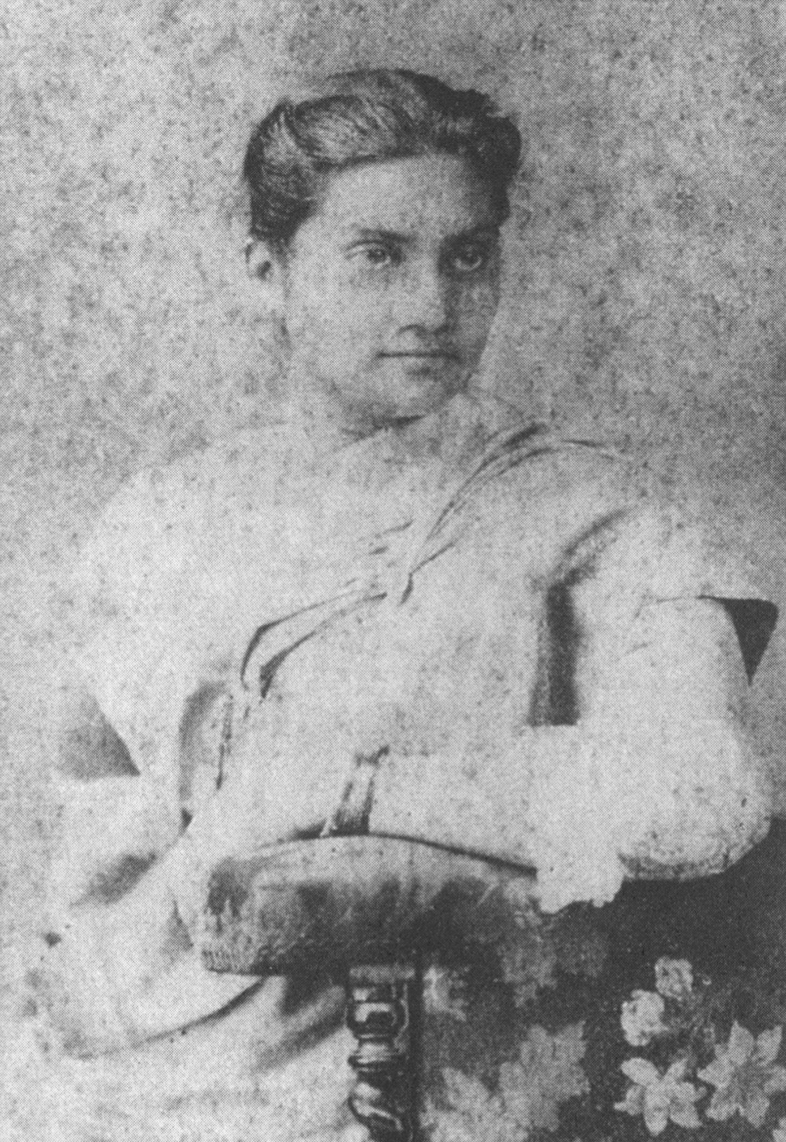 An Indian woman seen seated, dressed in white, resting her arm on the arm of a wooden chair. She looks half to the right.