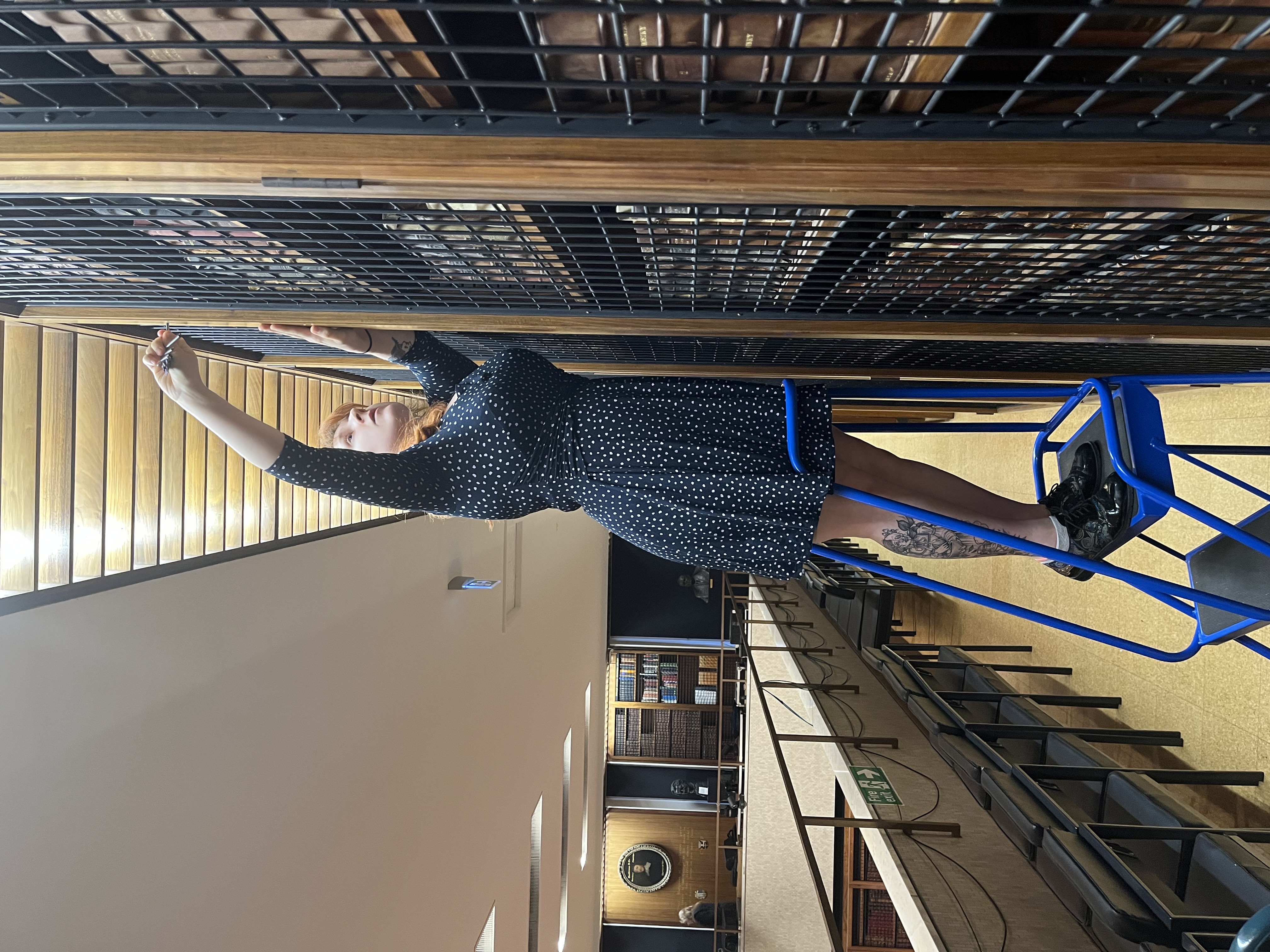 Image shows Kate, a white woman in her twenties, atop a blue library ladder. She is using a key to open a bookcase. She is on the first floor of the Royal College of Physicians Dorchester library. In the background, you can see more bookcases lining the walls of the room, and the portraits and busts of notable Royal College of Physicians members