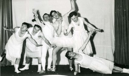 Black and white photograph of a group of men wearing white dresses performing on stage in a comedy show