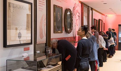 Photograph of men and women looking at an exhibition with objects in class display cases, and paintings and documents mounted on the walls.