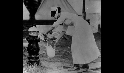 Black and white photograph of an influenza nurse wearing a face mask collecting water in a jug.