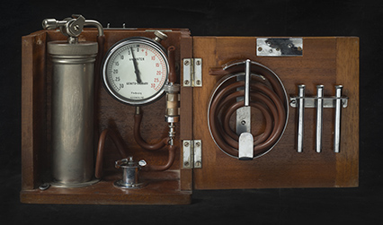 Wooden box containing a metal cylinder, pressure gauge, coil of rubber tubing and three small metal tubes.