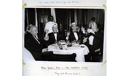 Black and white photograph of four men sat around a table, each wearing party hats