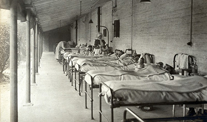 Black and white photograph of a row of hospital beds occupied by patients on a veranda. Nurses are attending to some of the beds.