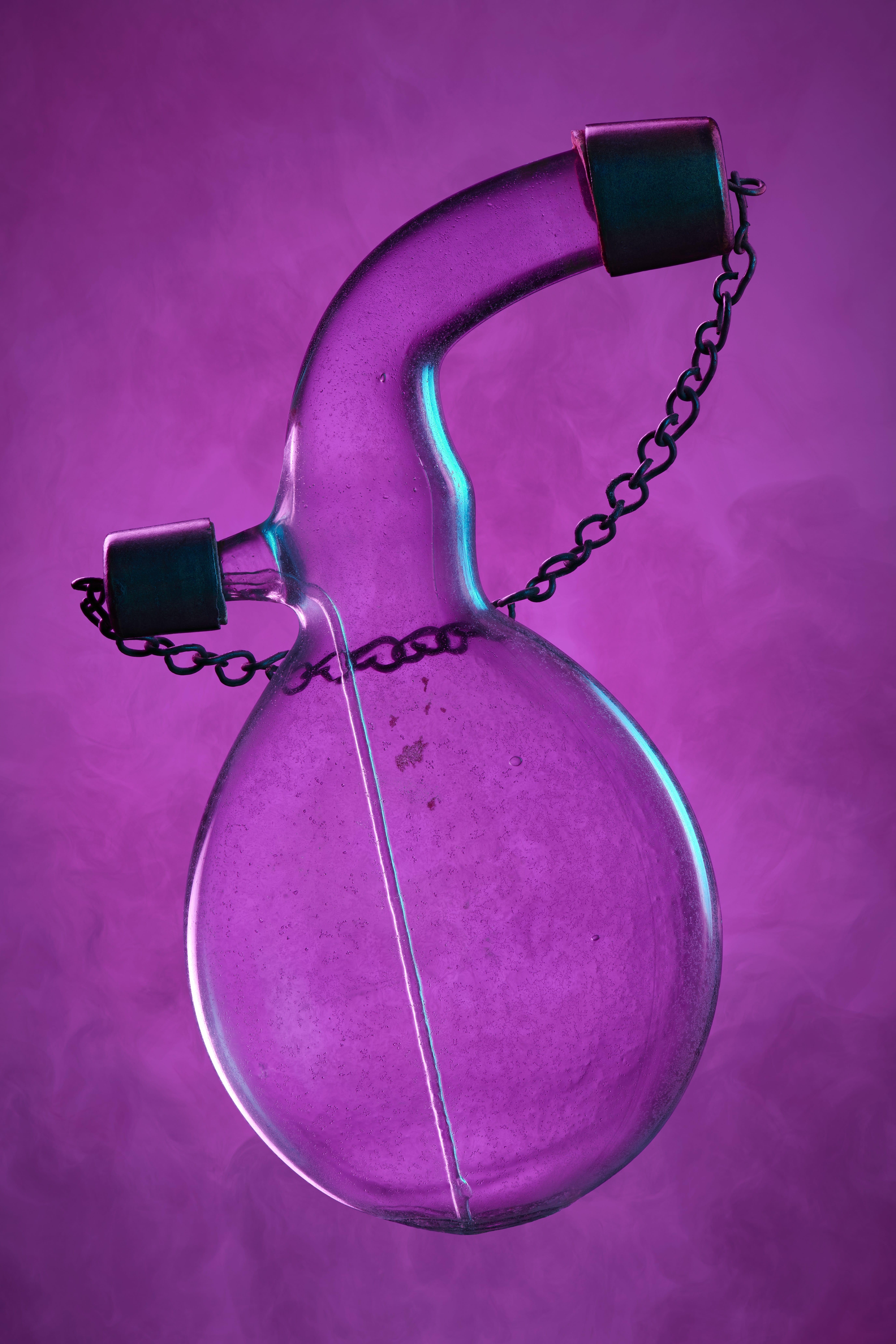 Circular glass flask with two necks, metal gap on each neck, caps connected by a small chain. All on a smoky pink background